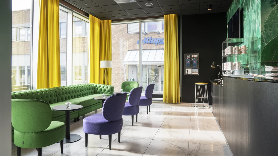 Lyst gulv, svart tak, grønn sofa, tre lilla stoler, en grønn stol, vinduer fra tak til gulv, gule gardiner, kaffestasjon med kaffekopper. 