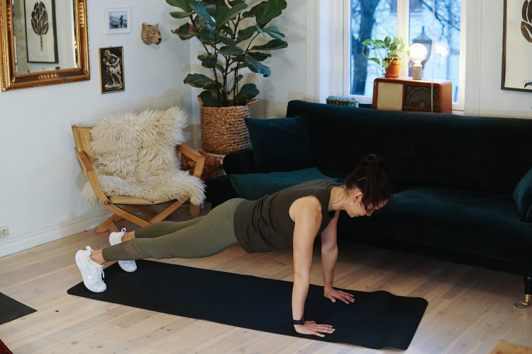 treningsekspert Anna Johansen trener på matte på gulvet, mørkegrønn sofa i bakgrunnen, lys stol med pels oppi, str grønn plante, bilder på veggen