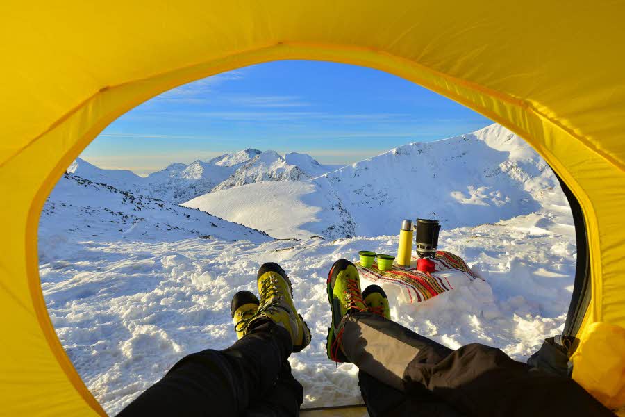 To par bein som kommer ut av en gul teltåpning i retning snødekte topper og leirplass.