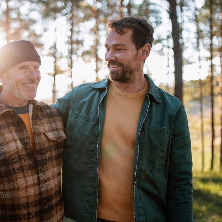 To smilende menn med rutete og grønn jakke går i skogen med trær og lysning i bakgrunnen