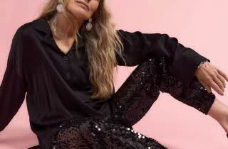 En kvinne sitter og poserer i sort skjorte og sort paljettbukse.