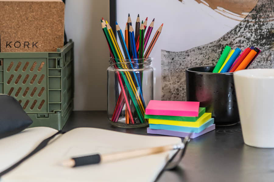 Nærbilde av et skrivebord med en åpen notatbok, blyanter i et glass og en bunke med fargede notatlapper