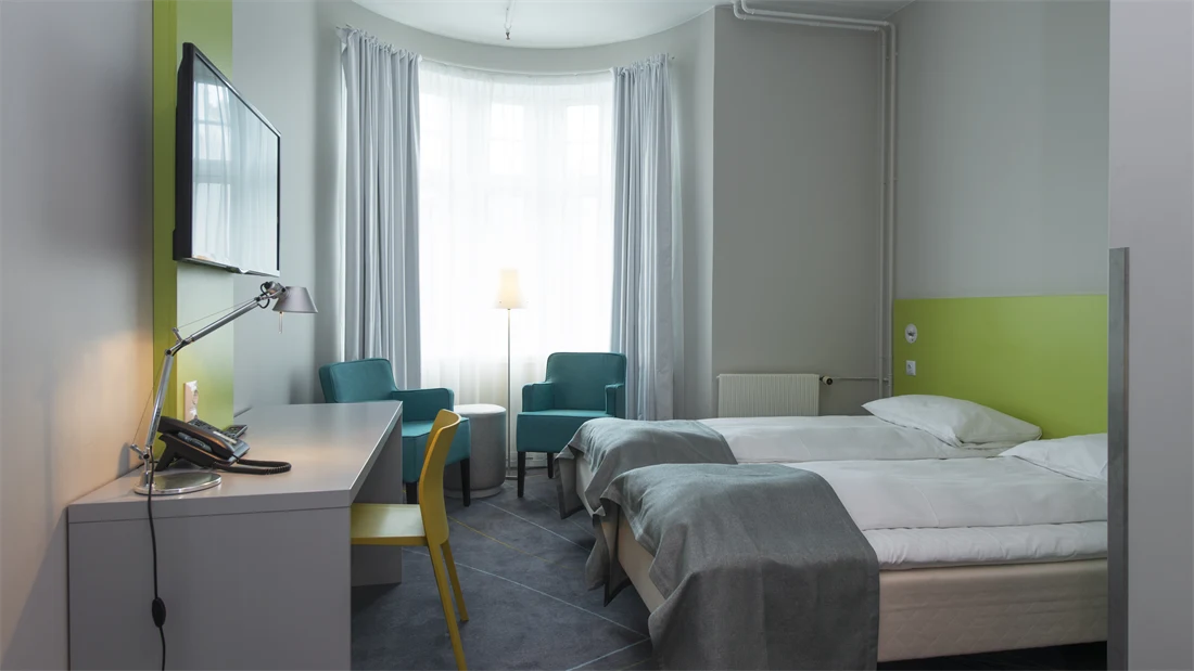 Standard Room Twin på Thon Hotel Trondheim. Lyst rom med store vinduer. Grått teppe, hvit vegg med limegrønne detaljer, to turkise lenestoler, to enkeltsenger, skrivebord med tilhørende gul stol. TV på veggen og stort speil.