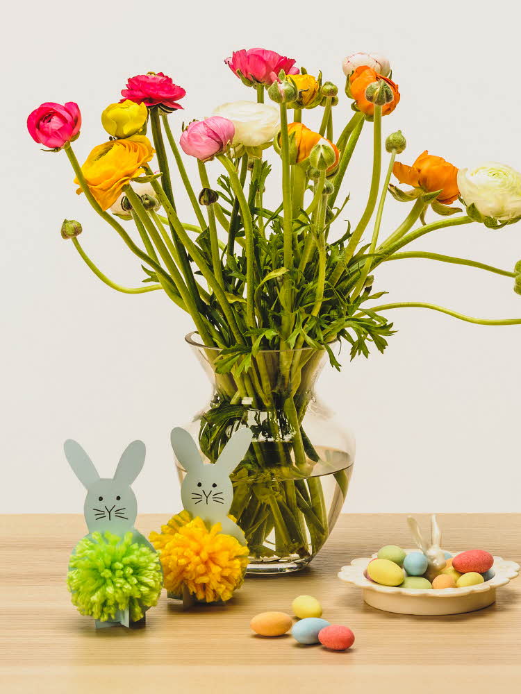 En gul og en grønn kanin laget av papir og garn ved siden av en saks  To grå kaniner med dusk laget av grønt og gult garn ved siden av en stor vase med blomster og sjokoladeegg på ett bord