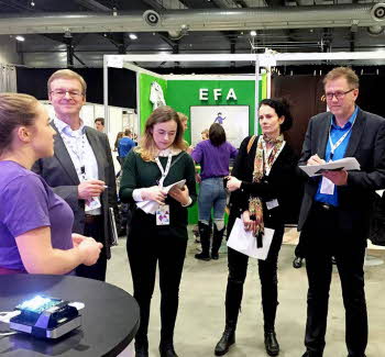 Fem medlemmer av en jury står i messehallen til Ungt Entreprenørskap og får presentasjon fra en ung gründer.