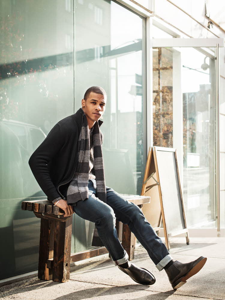 Mann med sort jakke, gråstripete skjerf, jeans og sorte chelseaboots sitter på benk foran glassfasade og ser i kamera