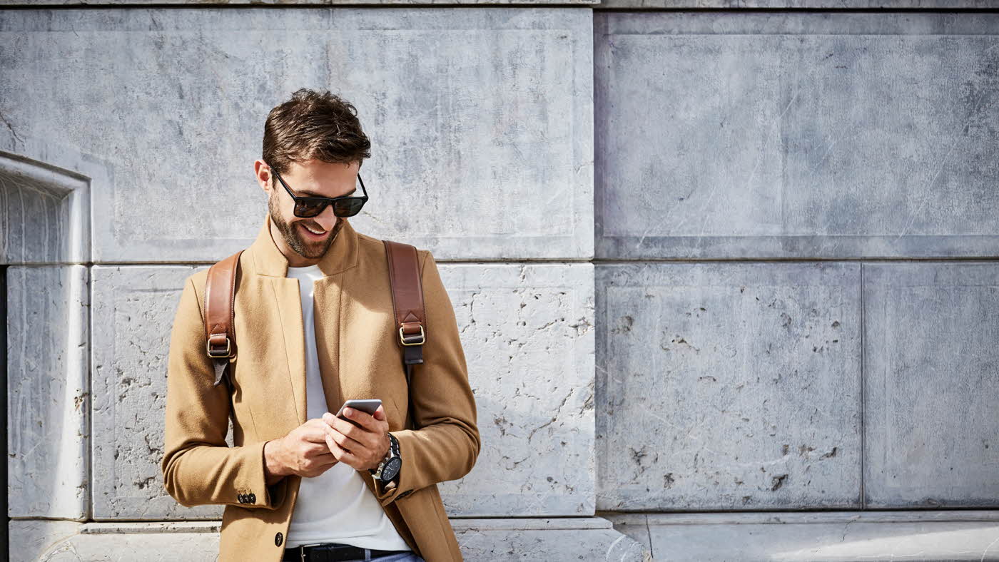 Mann med solbriller og lysebrun slim jakke ser på mobilen foran murvegg