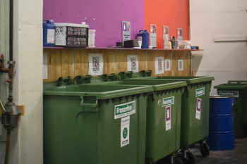 Godt organisert avfallsrom med grønne avfallsdunker og fargekoder og gjenstander på veggen som forklarer hva som kan kastes i disse avfallsdunkene.