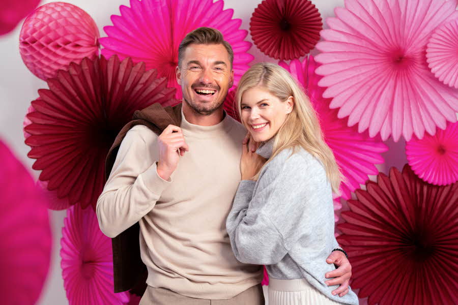 Mann som holder rundt en dame mens begge smiler til kamera med rosa papirdekor i bakgrunn