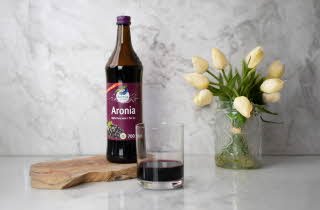 Aronia-juiceflaske og et glass juice på en treplate med tulipaner i bakgrunnen.