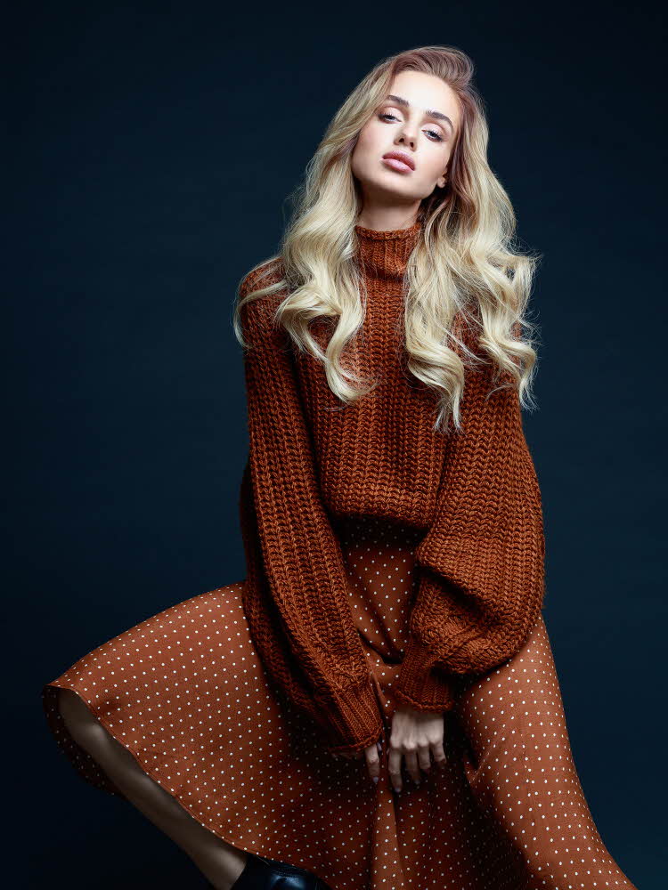 Dame med brun ullgenser og brun- og hvitprikkede skjørt poserer på stol