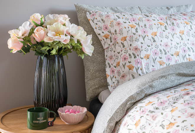 Seng med blomstrete sengetøy og nattbord med blomster og en kopp.