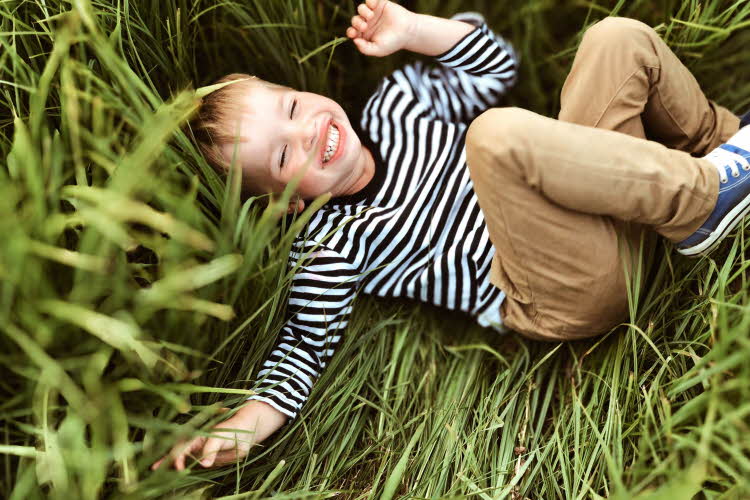 Gutt ler og ligger i gresset iført stripete genser og beige bukse