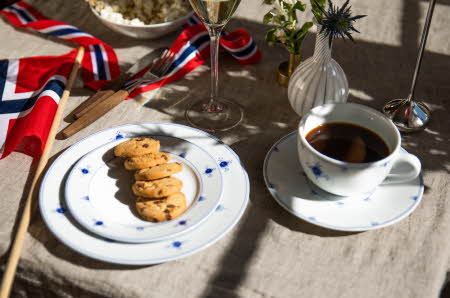 Et bord ute dekket med et hvitt servise med blå blomster, og et norsk flagg liggende ved siden av. Det er kjeks på asjetten og kaffe i koppen.