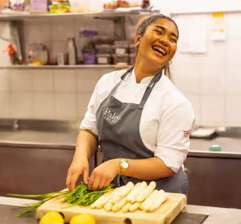 Kokken Mymint på Thon Hotel Lofoten står i et industrielt kjøkken og skjærer opp asparges, smiler og ser opp. Benk, vegg og hylle i bakgrunnen er utvidet med kunstig intelligens for å gjøre bildeformatet større enn opprinnelig.