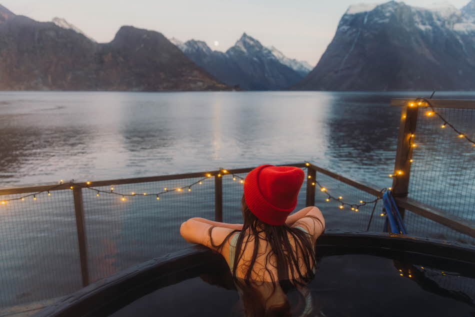 Dame med rød lue sett bakfra sittende i badestamp med fjord- og fjellandskap.