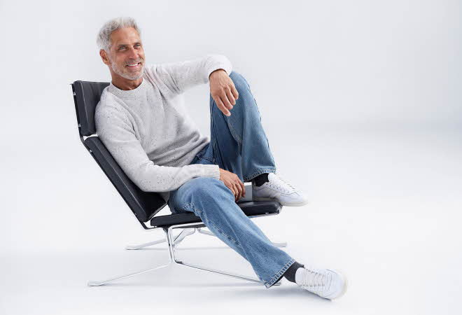 En mann i grå genser og jeans sitter i en stol og smiler mot kamera.