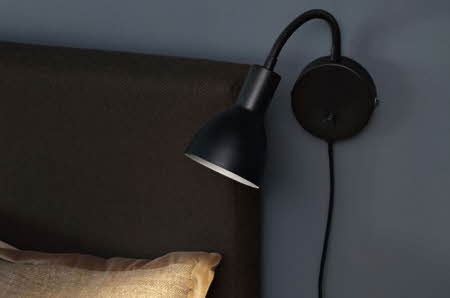 Sort vegglampe med fleksibel arm montert på mørk vegg ved siden av en sengegavl.