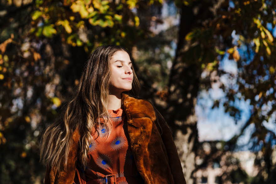 Dame med brunt hår, brun fuskepelskåpe og brun og blå silkebluse lukker øynene i solen foran høsttrær