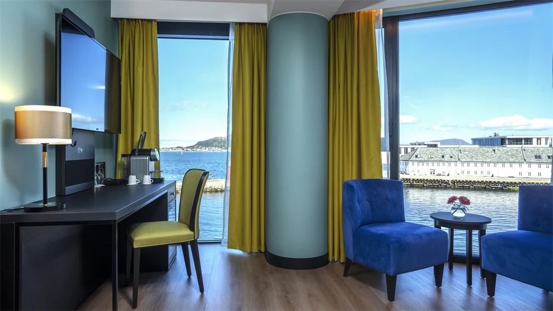 Gule gardiner, panoramavindu med utsikt over vannet, to blå lenestoler, salongbord, turkis vegg, skrivebord