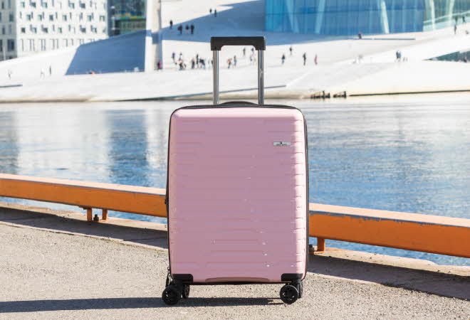 Rosa hardplastkoffert med fire hjul ved vannkanten.