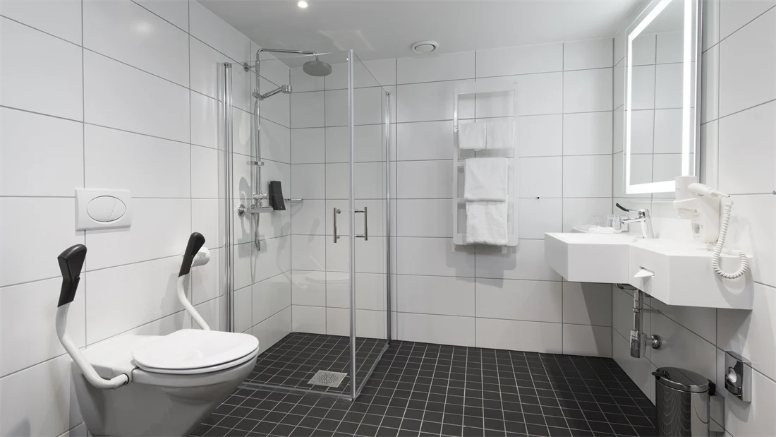 Toalett med støttearmer, mye gulvplass, dusj, vask og speil på bad i handikaprom på Thon Hotel Rosenkrantz Oslo, som ligger i Oslo sentrum