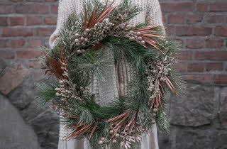 Person holder en stor dekorativ julekrans med grønne og bronsefargede grener foran en murvegg.