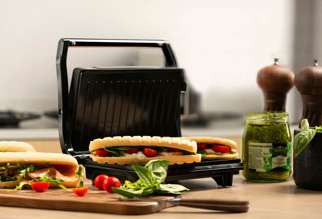 En sandwichgrill med to varme sandwicher, omgitt av ferske ingredienser på kjøkkenbenken.