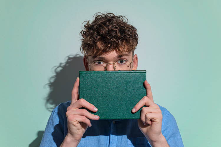 Gutt med briller og blå skjorte holder grønn bok foran nedre del av ansiktet sitt