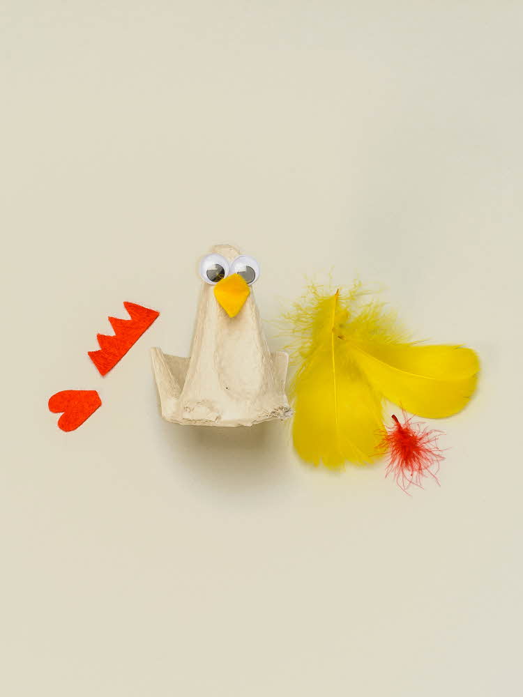 En eggekartong omgjort til en høne med øyne og nebb, resten av utstyret ligger ved siden av  En eggekartong omgjort til en høne som mangler vinger og hanekam