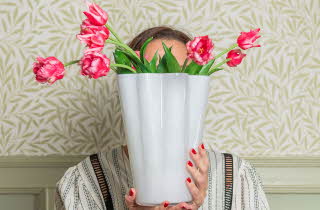 Dame som holder en vase med rosa blomster foran ansiktet.