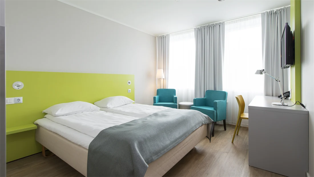 Standard Room Double på Thon Hotel Trondheim. Dobbeltseng med grått sengeteppe. Limegrønn sengegavl. Grått skrivebord med tilhørende gul stol. To turkise lenestoler og et rundt bord. 