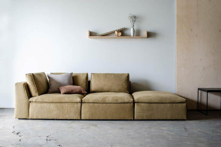 Beige stor sofa, i et hvitt rom med trefiner på en liten del av veggen. Hylle i tre på veggen med en vase med strå i. et lite svart bord helt til høyre i bildet. 