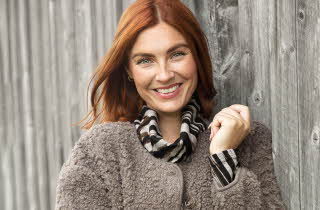 En kvinne i brun jakke står og lener seg mot en vegg og smiler