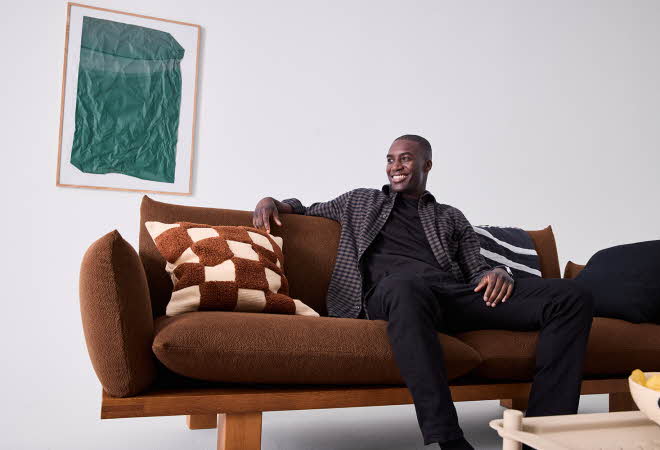 En mann som sitter på en brun sofa med mønstrede puter.
