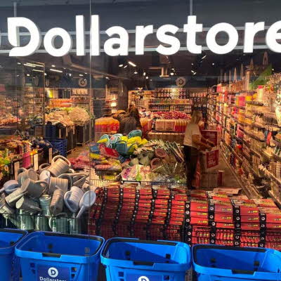 Glassvegg av butikken til Dollarstore med kunder som handler i butikken, blå handlekurver og et stort lys skilt av Dollarstore sin logo