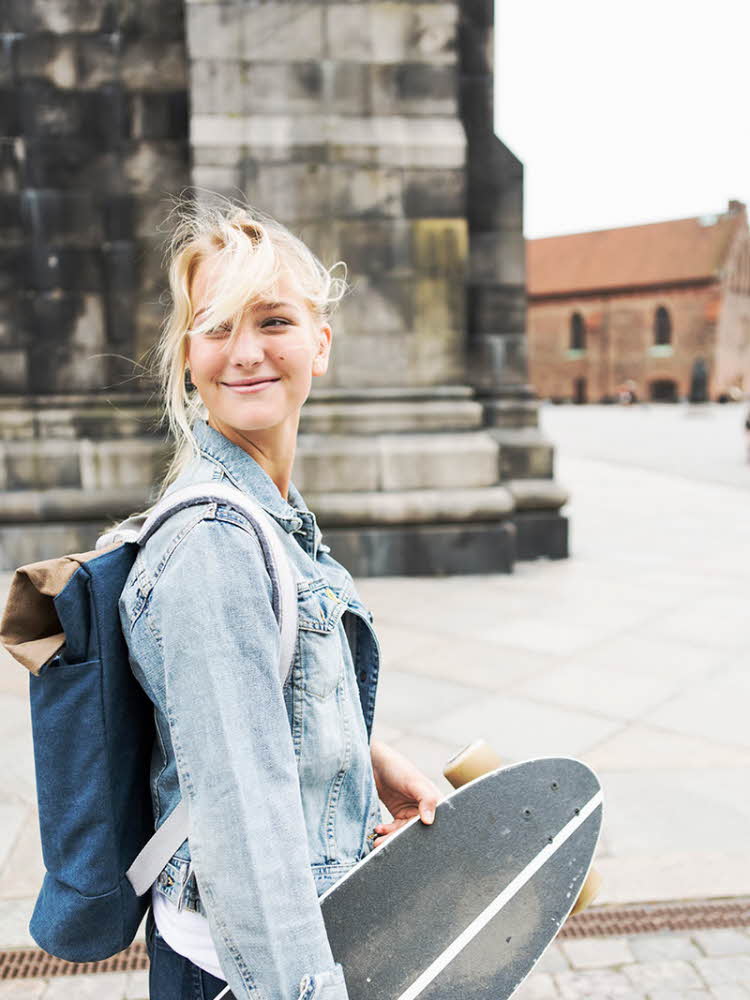 Smilende blond jente som går med sekk på ryggen og skateboard under armen