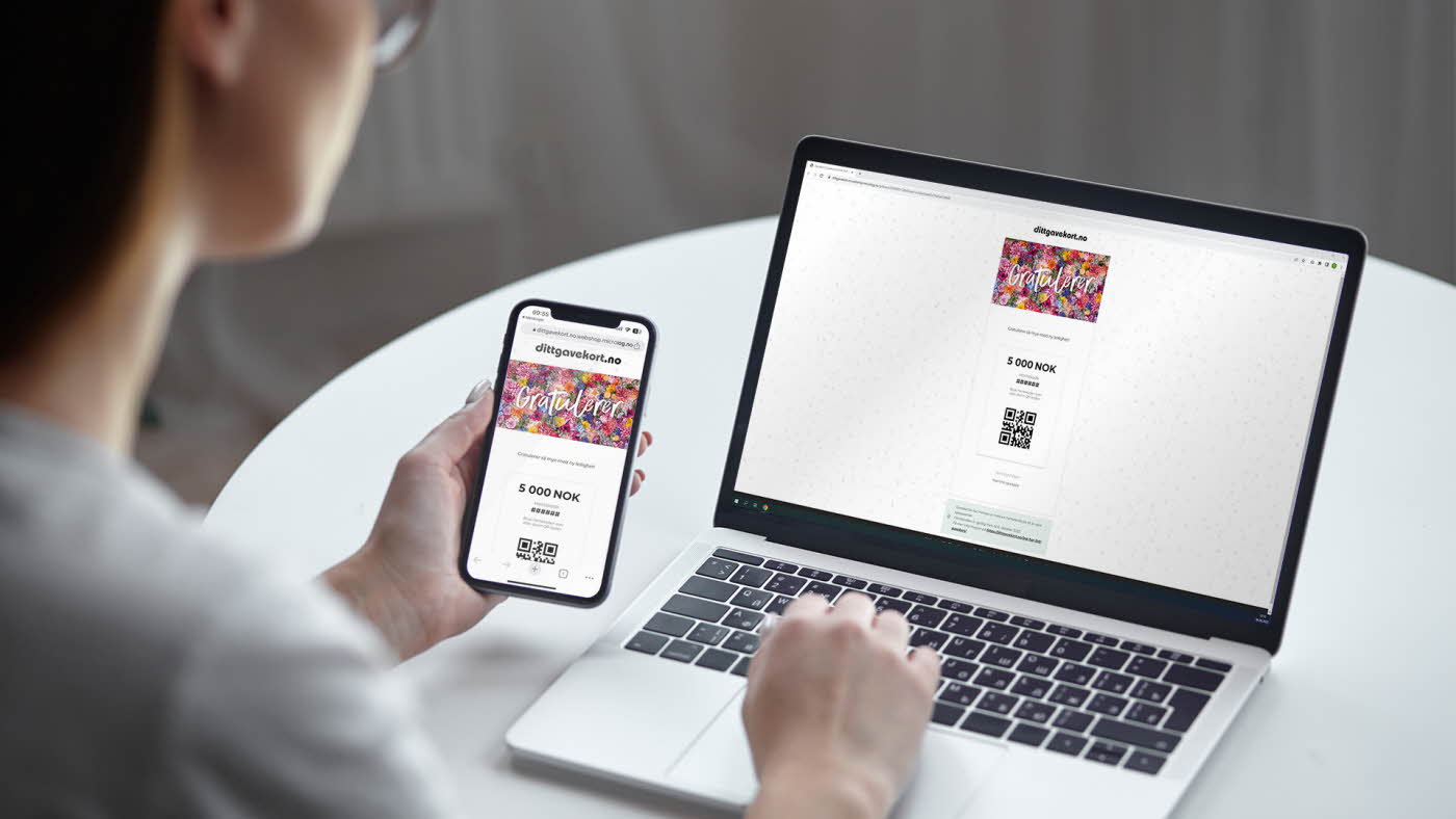 Perspektiv fra overskulderen på person som sitter ved et hvitt, rundt bord med en PC som viser bilde av digitalt gavekort fra dittgavekort.no og personen holder også en mobil med samme motv som på PC-en.