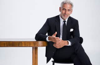 Mann i svart dress og hvit skjorte sittende ved et bord.