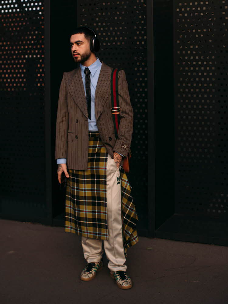 Herremodell utendørs i brun blazer, blå skjorte, slips, rutete skjørt, beige chinos og sneakers.