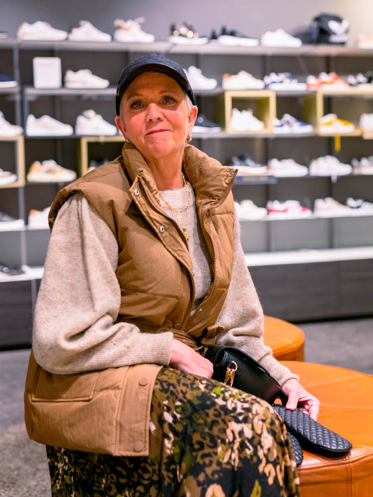 dame i mønstrete kjole, lysegrå strikkegenser over og beige vest utenpå. mørkt hodeplagg og mørk veske