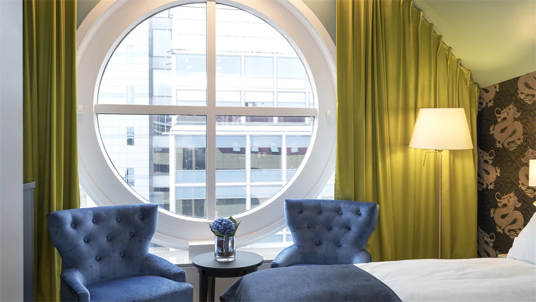 Rundt vindu med utsikt, dobbeltseng og blå lenestoler og gule gardiner på Thon hotel Terminus i Oslo sentrum nær Jernbanetorget