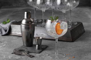 Et cocktailglass med is, appelsinskive og mynte står i sentrum, omgitt av shaker, målebeger og tomme krystallglass.