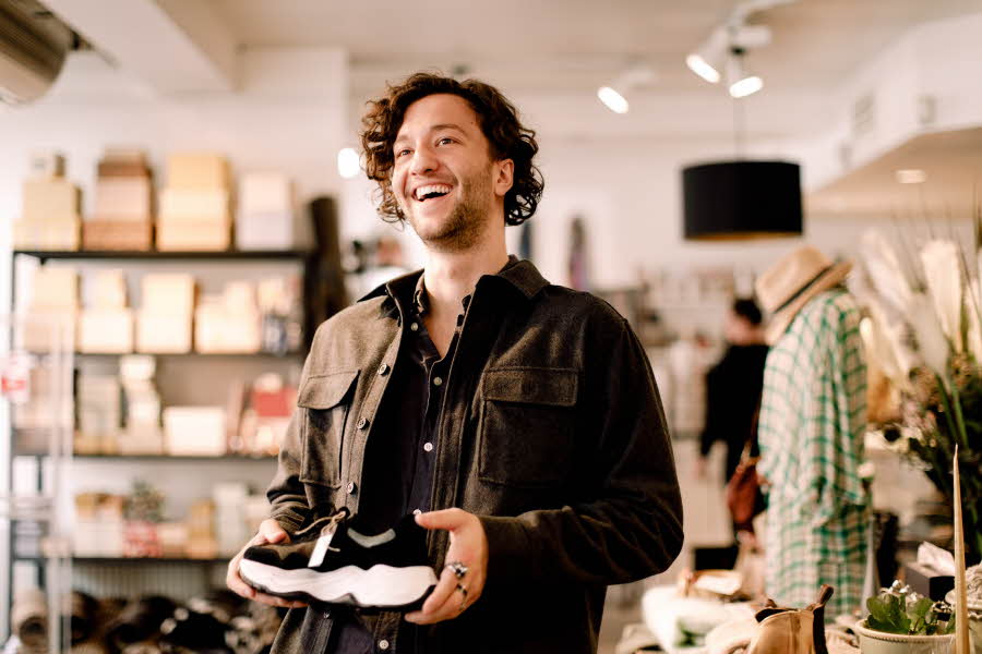Mann på shopping med mørkt krøllete hår og brun jakke holder en sko i hendene inne i en klesbutikk