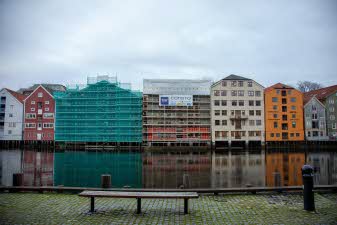 Utsnitt av Kjøpmannsgata som speiler seg i Nidelva, med nr. 15 i midten. I forgrunnen en benk.