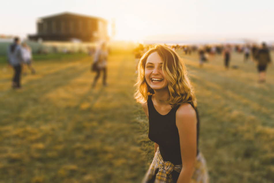 Jente med sort topp og gul rutete skjorte knytt rundt livet smiler til kamera med festival i bakgrunnen