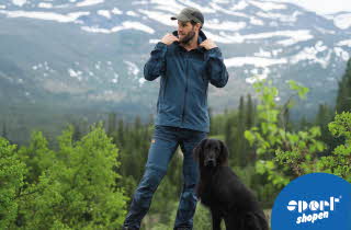 Mann i blå turklær med svart hund foran snødekte fjell.