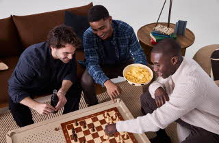 Tre menn som spiller sjakk ved et stuebord.