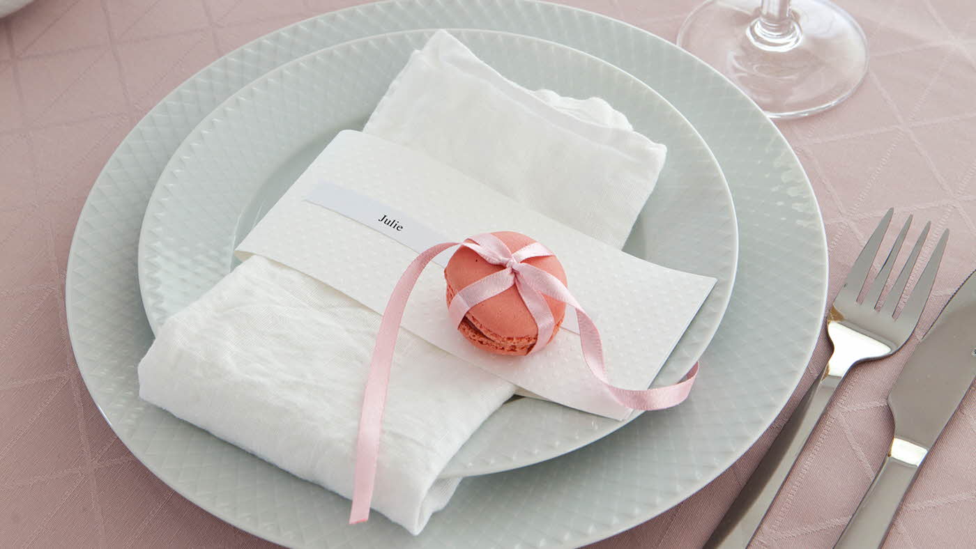 To hvite tallerker, kniv og gaffel, rosa duk og hvit serviett med rosa makron knyttet med silkebånd og en navnelapp hvor de står Julie