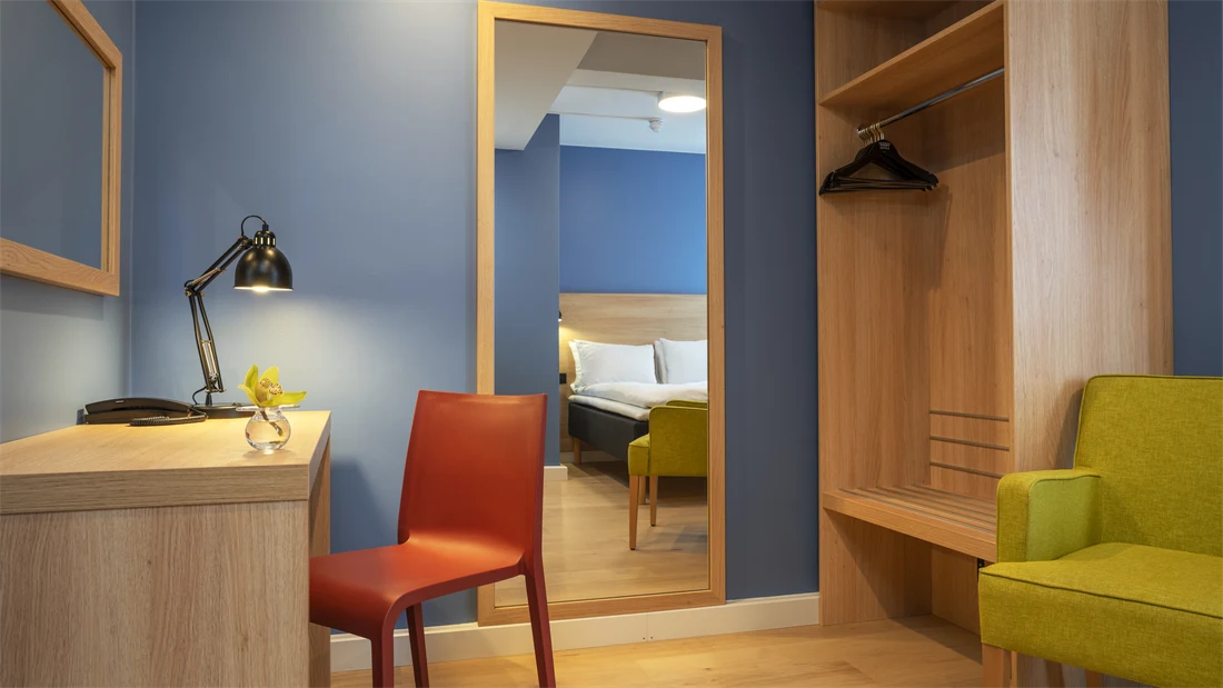 Skrivebord, speil, blå vegger, garderobe og gul stol i standard rom queen på Thon Hotel Astoria